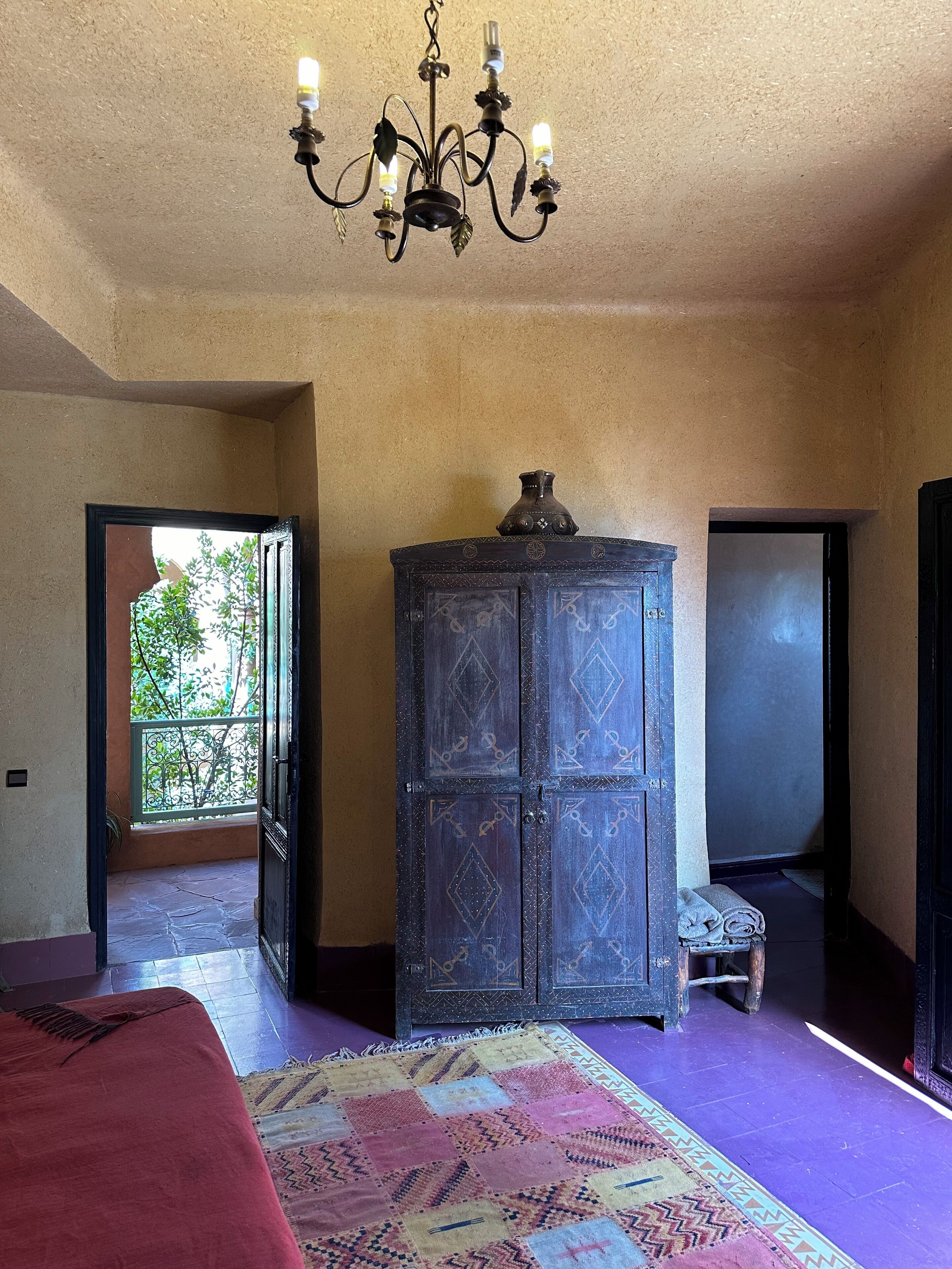 Una habitación con una araña, un armario grande, una cama con una manta roja y una alfombra colorida.
