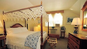 Un lit à baldaquin avec des poteaux aux quatre coins, un ciel de lit blanc, une tête de lit en bois et une couvre-lit à motifs bleus et blancs. On y trouve une table de chevet en bois avec une lampe, une commode avec un miroir et une petite table avec une lampe. Une télévision ancienne est posée sur un meuble à côté du lit.