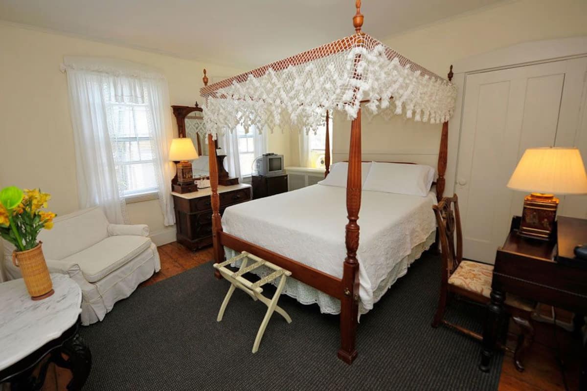 Una cama con dosel y cortinas, una mesita de noche con lámpara, una pequeña silla, un buró con lámpara y un jarrón con flores.