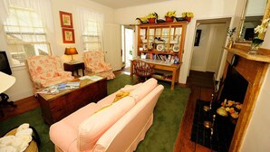 Un salon chaleureux avec un canapé rose, des fauteuils à motifs floraux, une table basse en bois et une bibliothèque remplie de livres et d’objets décoratifs.