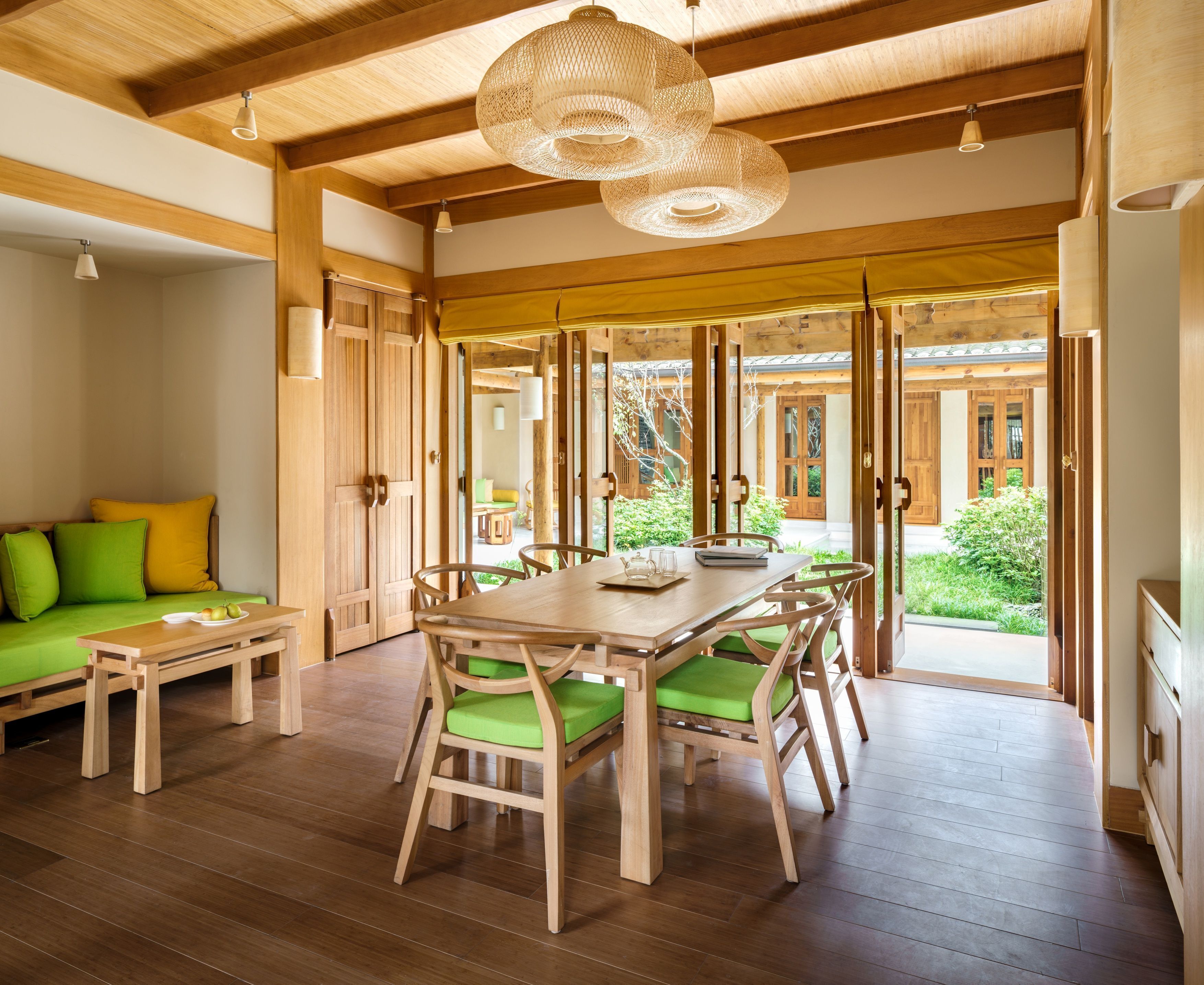 A spacious room with a wooden dining table, chairs, and a bench. There is a sofa with cushions and a small table with a bowl of fruit. The room has wooden floors and a large window with a view of greenery outside.