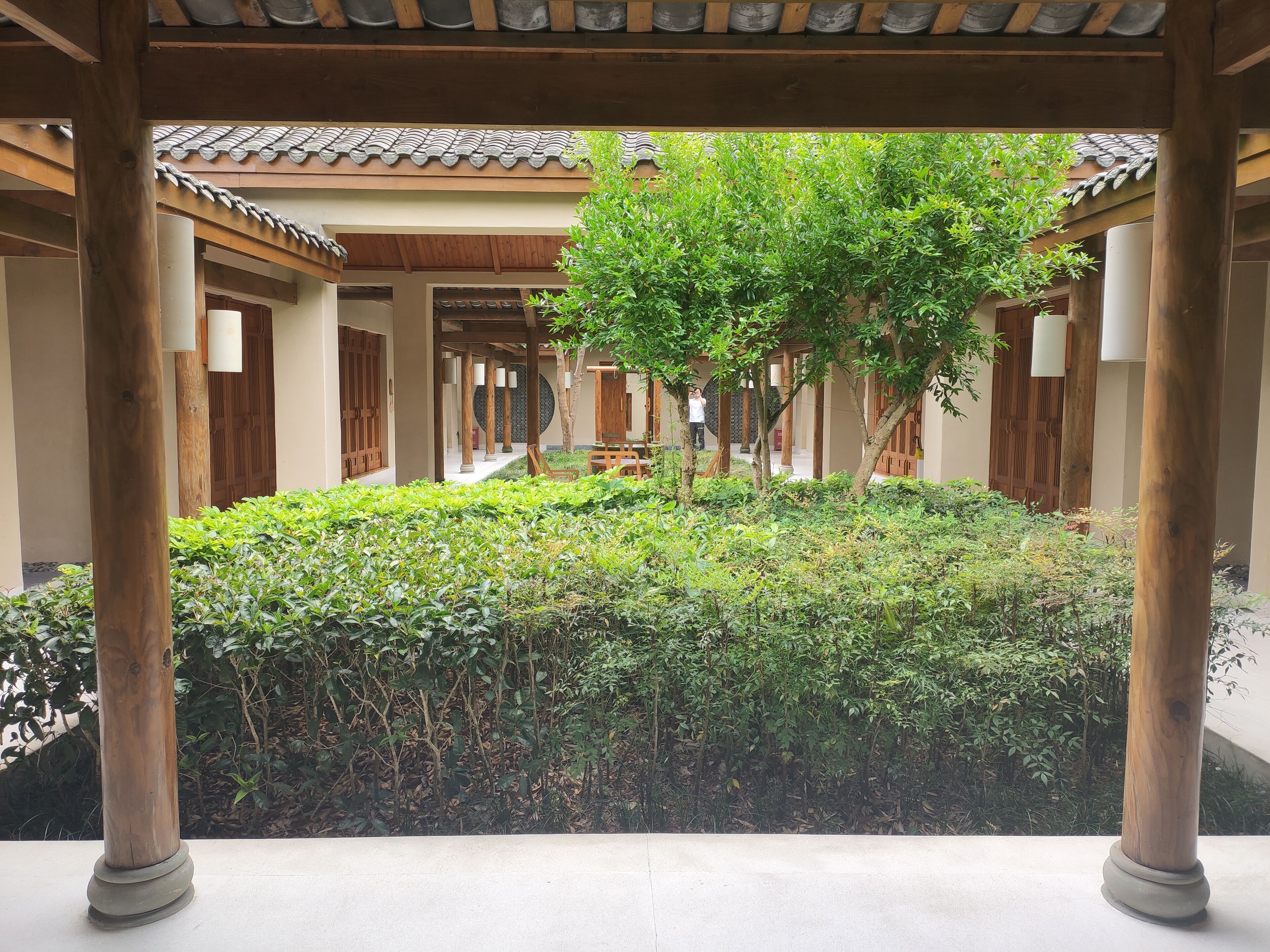 A traditional wooden pavilion with a tiled roof, surrounded by lush greenery and a small tree.
