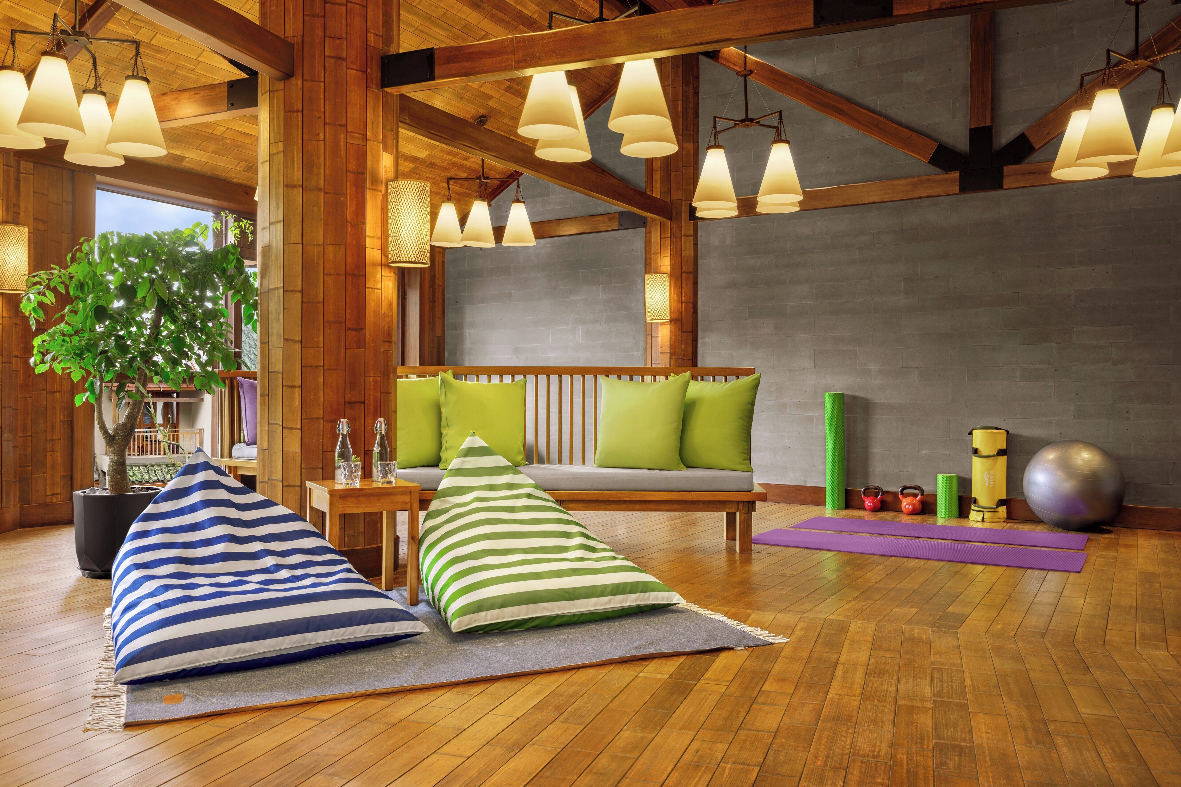 Um quarto espaçoso com vigas de madeira, uma janela grande, um sofá com almofadas verdes, um tapete de yoga e equipamentos de ginástica.