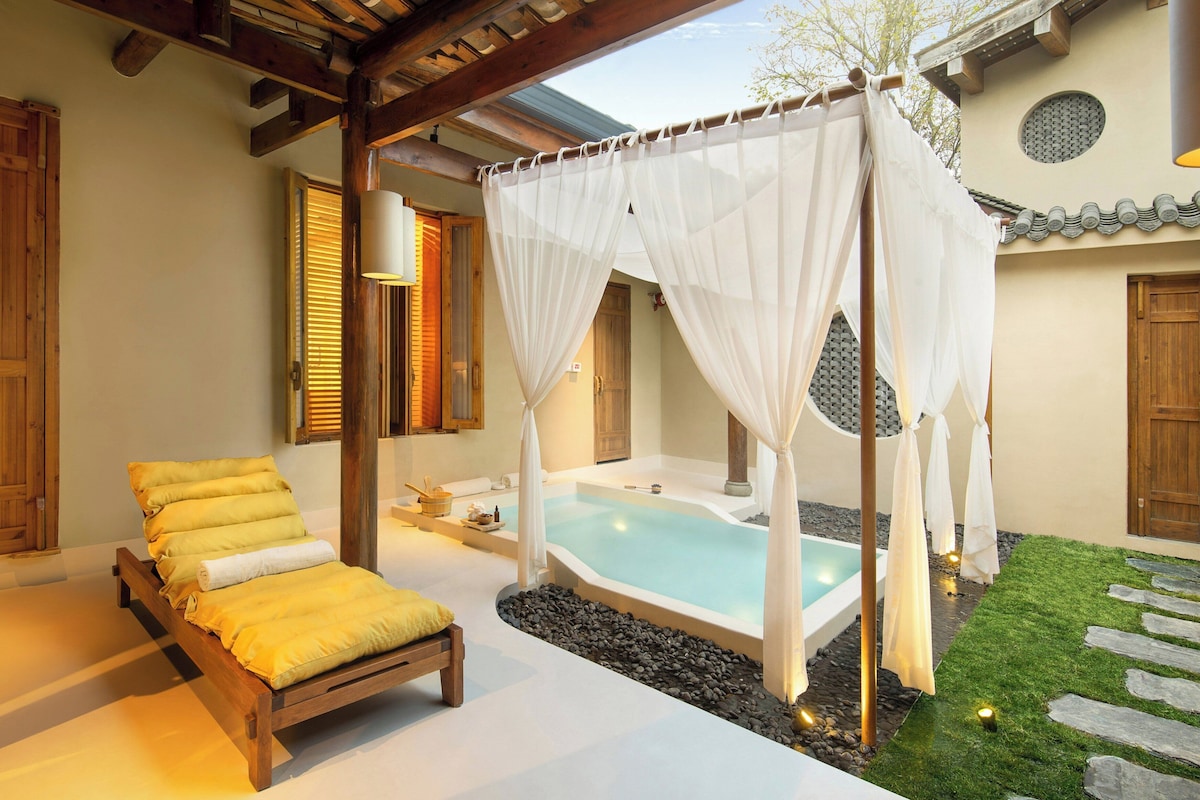 Une chambre avec un lit en bois, un petit coin salon, un jacuzzi extérieur privé et une pergola agrémentée de rideaux blancs.