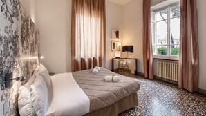 Habitación de hotel con cama, escritorio, silla, radiador y ventana con cortinas.