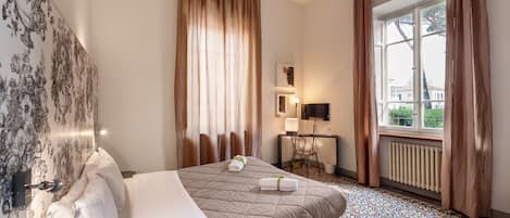 Habitación de hotel con cama, escritorio, silla, radiador y ventana con cortinas.
