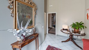 Una habitación con un espejo grande y ornamentado, una consola de madera con objetos decorativos, una mesa redonda pequeña con una lámpara y una planta en maceta, y una silla roja.