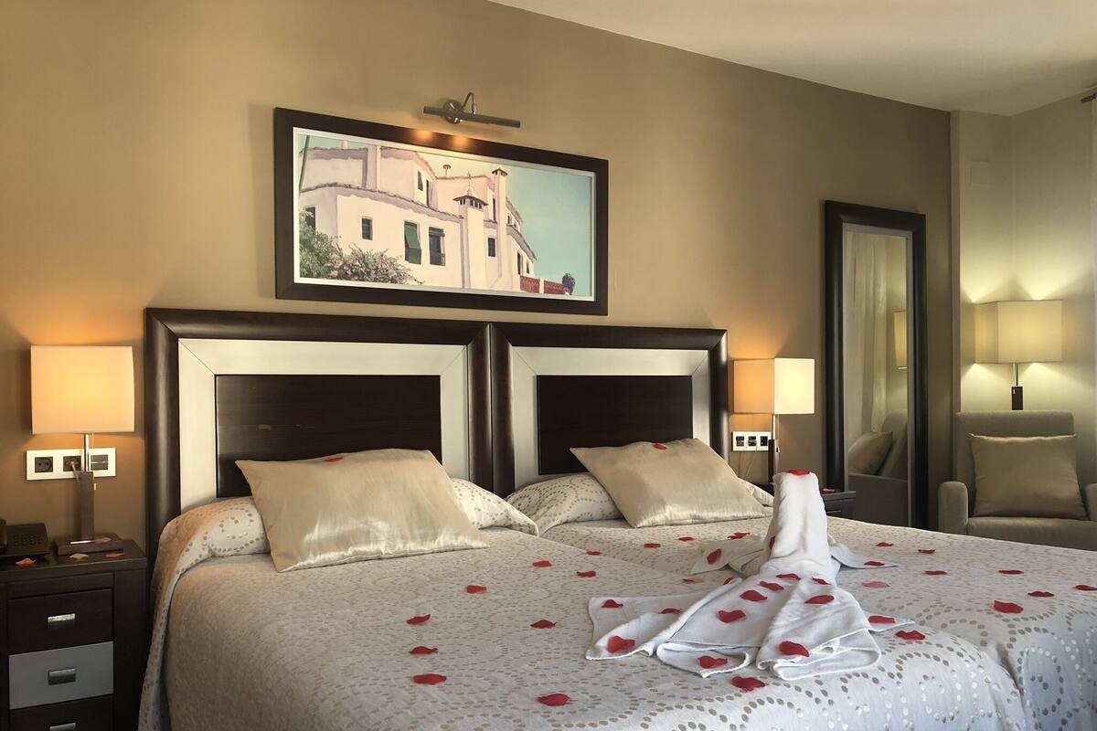 A double bed with a decorative towel and rose petals, a painting above, and two bedside lamps.