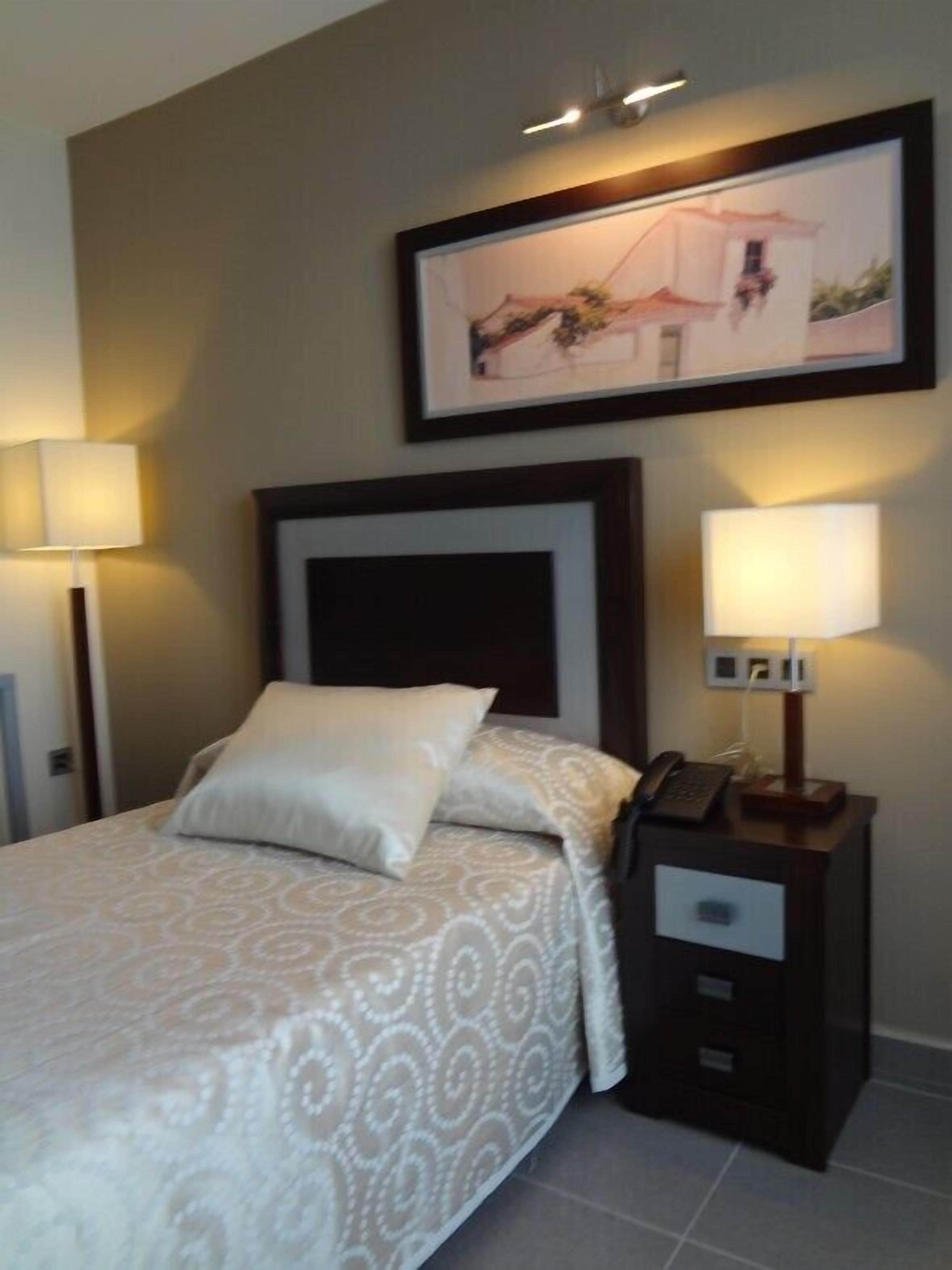 Un lit bien fait, avec une couvre-lit à motifs et des oreillers, une table de chevet avec une lampe, et un tableau encadré au mur.