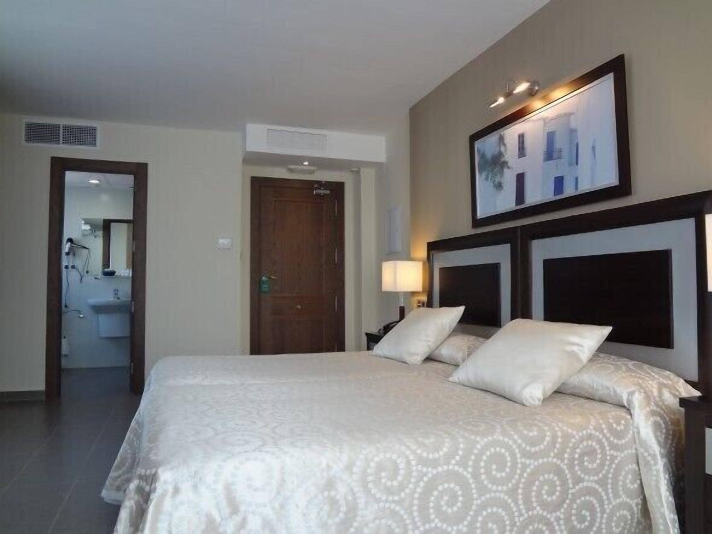 Habitación de hotel con una cama grande, una mesita de noche con lámpara, un cuadro en la pared y una puerta que da al baño.