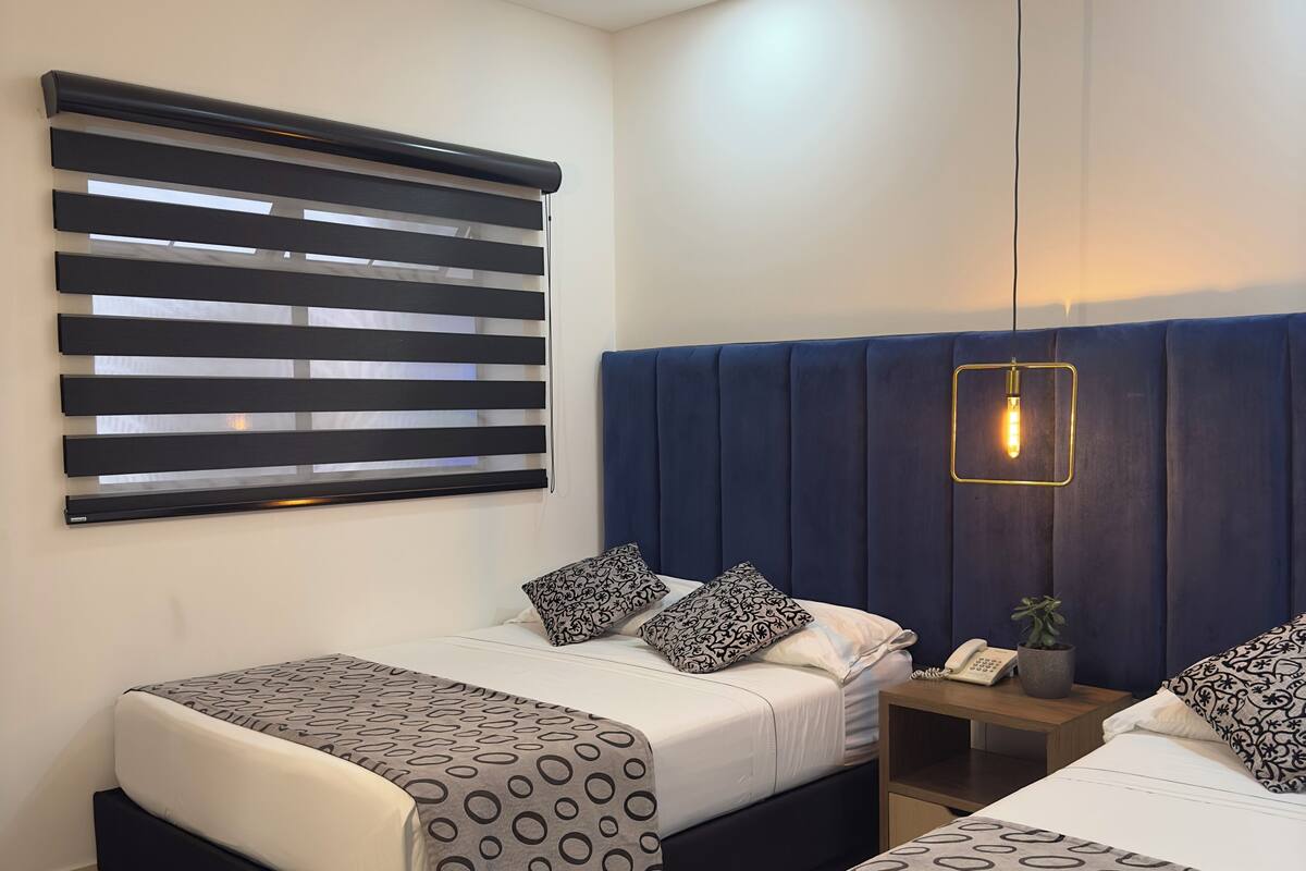 Una habitación de hotel moderna con dos camas, un ventanal con persianas, una mesita de noche con lámpara, y una mesita con teléfono y planta.