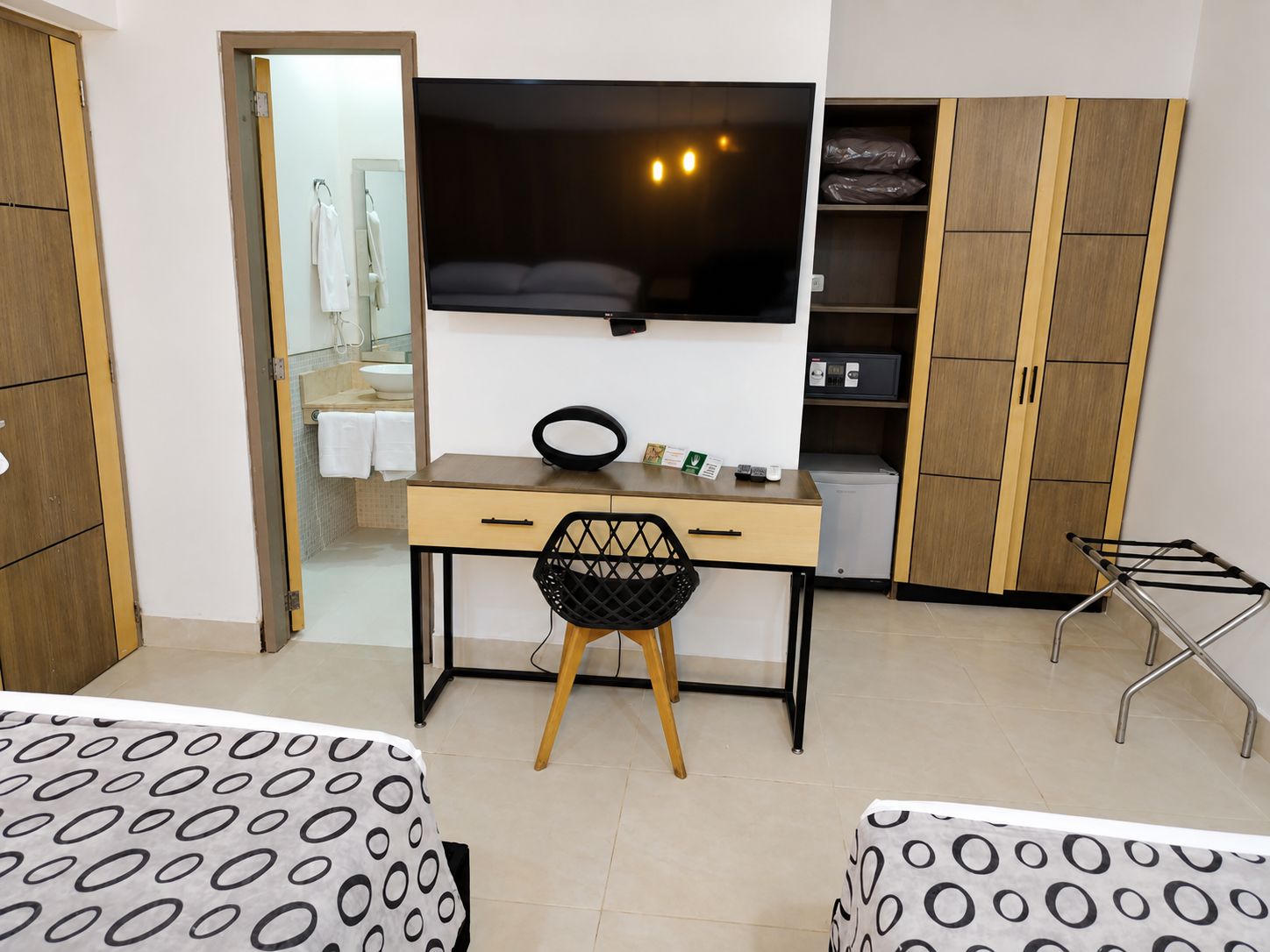 A hotel room with a bed, a desk with a chair, a TV, a wardrobe, and a small kitchenette.