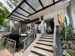 Entrada de un hotel con una escalera de cristal, un área cubierta con vigas de metal y varias banderas.