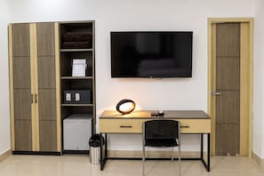 Une chambre avec un téléviseur fixé au mur, un bureau avec une chaise, un petit réfrigérateur, une poubelle et une armoire avec un miroir.