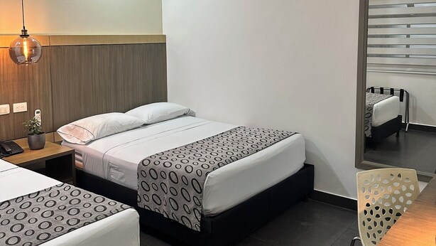 A hotel room with two beds, a desk, a chair, a mirror, an air conditioning unit, and a hanging light fixture. - Hotel Merlott Lleras (Medellín)