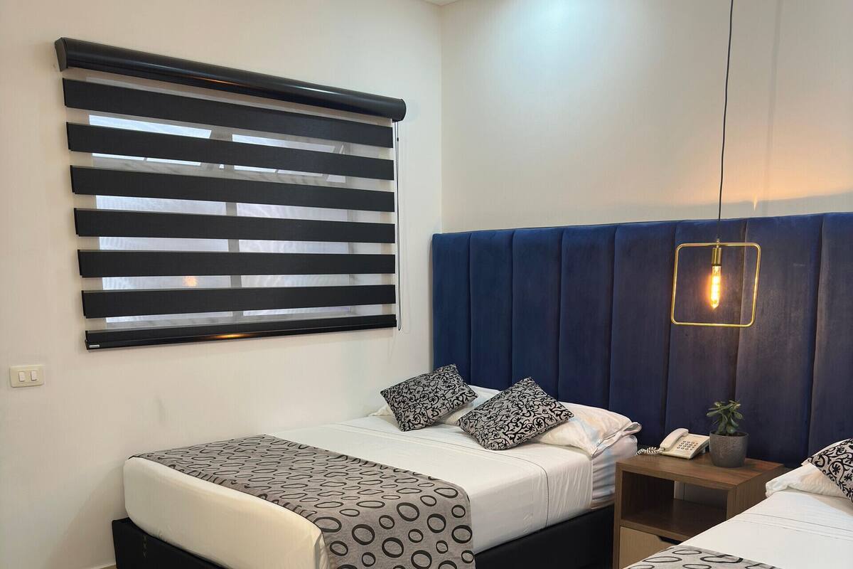 Una habitación de hotel moderna con dos camas, un ventanal con persianas, cabecera azul, mesita de noche con lámpara y una planta pequeña.