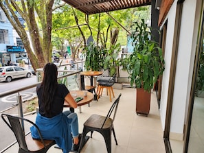 Une femme est assise à la terrasse d’un café, d’où elle a une vue sur la rue et la verdure.