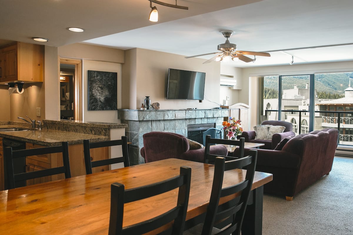 A modern kitchen with granite countertops, a dining area with wooden tables and chairs, a fireplace, and a large window offering a view of the mountains.