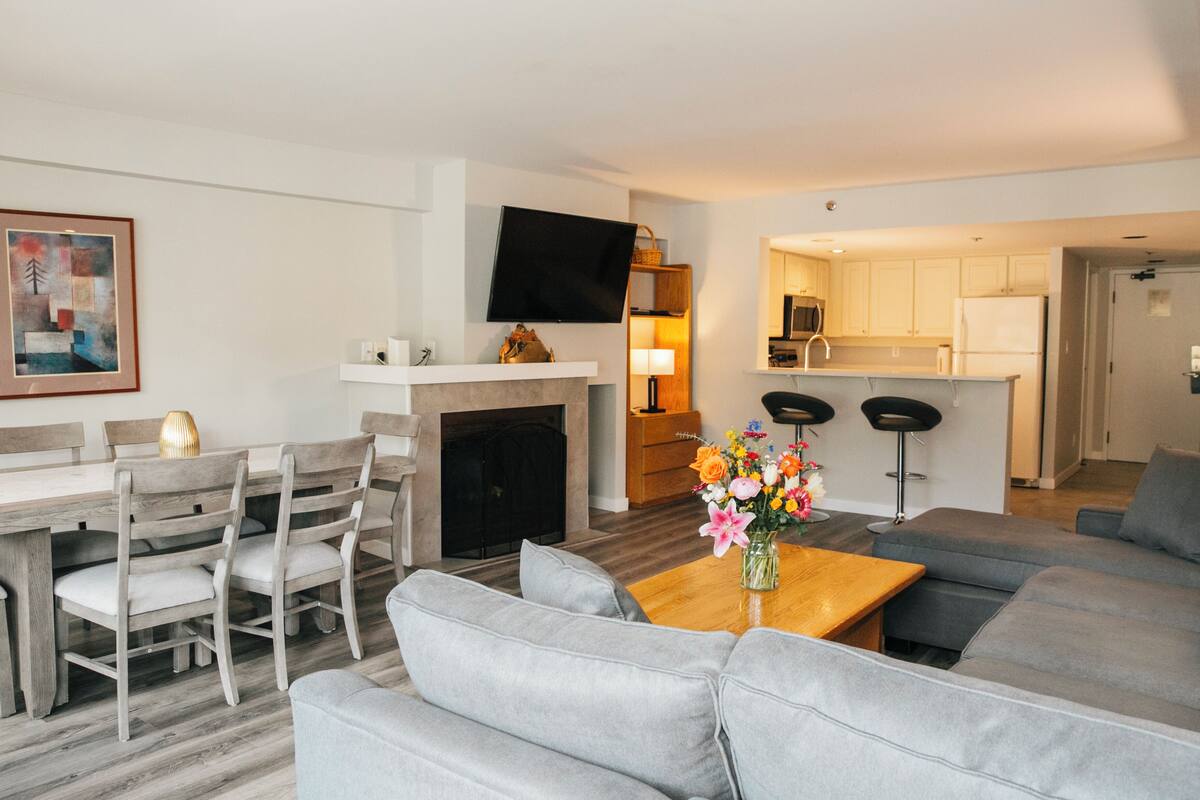 A modern living room with a grey sofa, a wooden coffee table with a vase of flowers, a dining area with a wooden table and chairs, a flat-screen TV mounted above the fireplace, and a kitchen area with white cabinets and a microwave.