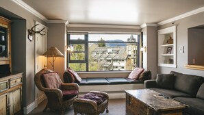 A cozy room with a built-in bench, wicker furniture, a wooden coffee table, and a view of buildings through a large window. - Carleton Lodge (Whistler)
