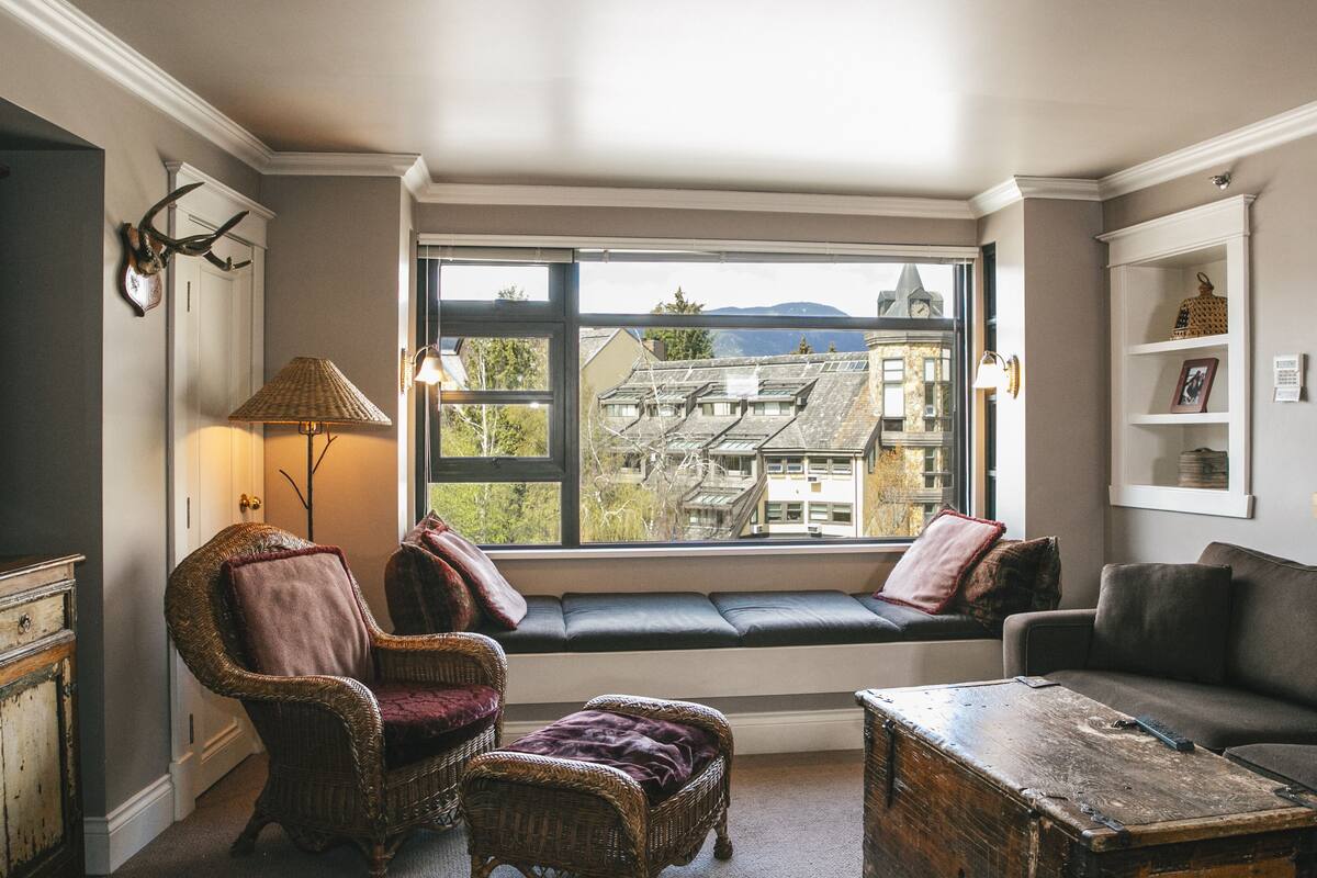 A cozy room with a built-in bench, wicker furniture, a wooden coffee table, and a view of buildings through a large window.