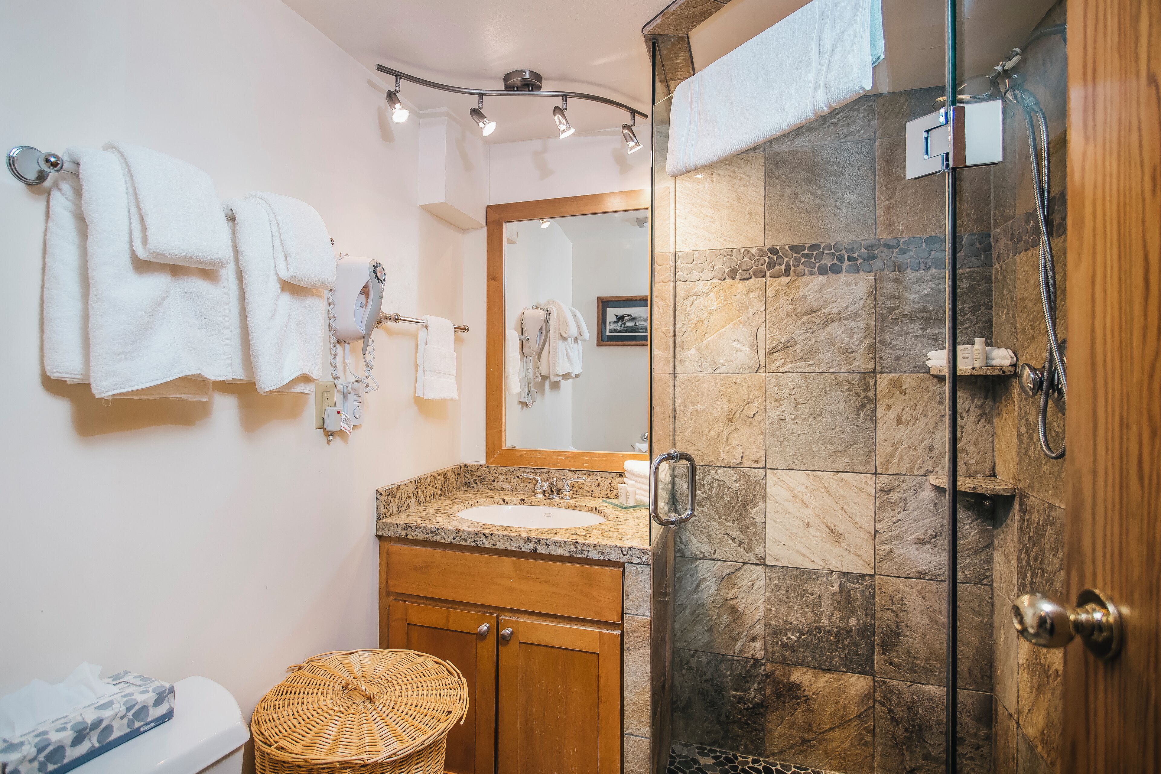 A bathroom with a granite countertop sink, wooden cabinets, a wicker basket, a glass-enclosed shower, and multiple white towels hanging on hooks.