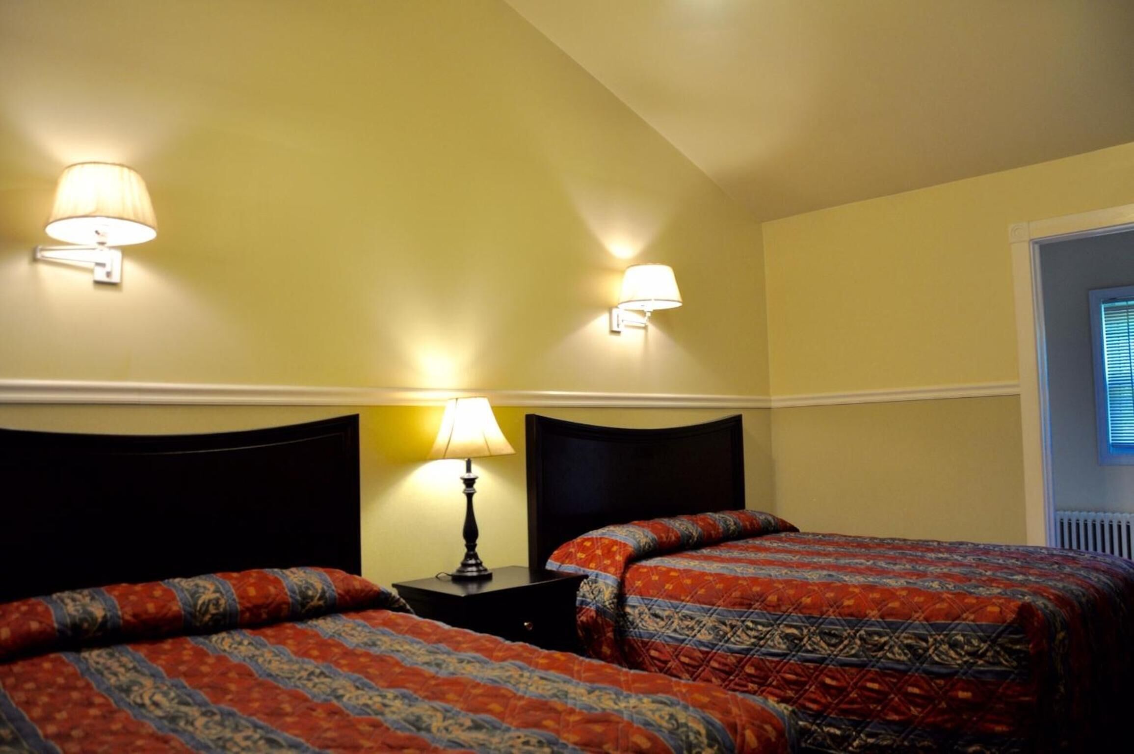 Quarto de hotel com duas camas, cada uma com roupa de cama estampada, uma mesa de cabeceira com abajur e luminárias na parede.