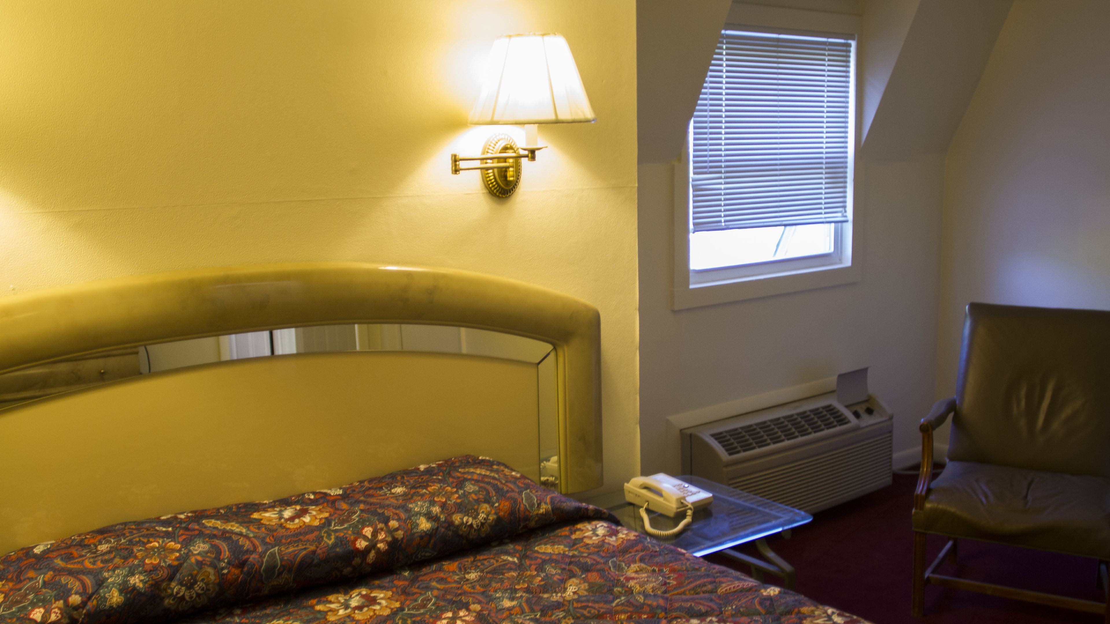 A hotel room with a bed, a chair, a lamp, a phone, and a window with blinds.
