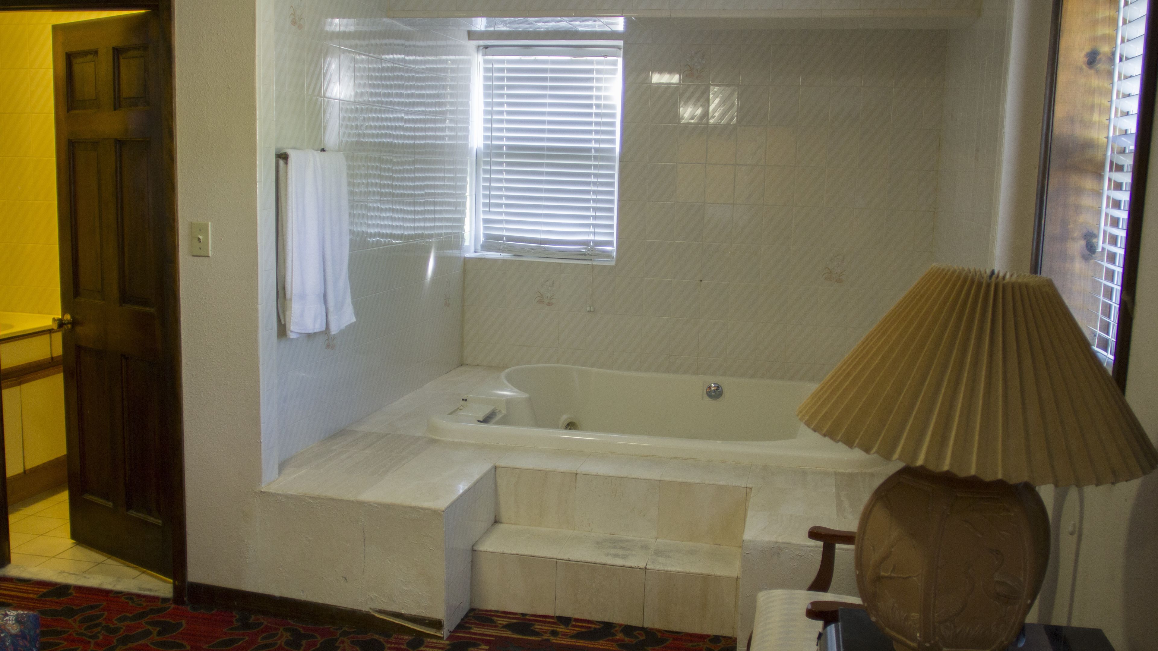 Un baño con bañera, una ventana con persianas, un perchero para toallas y una lámpara.