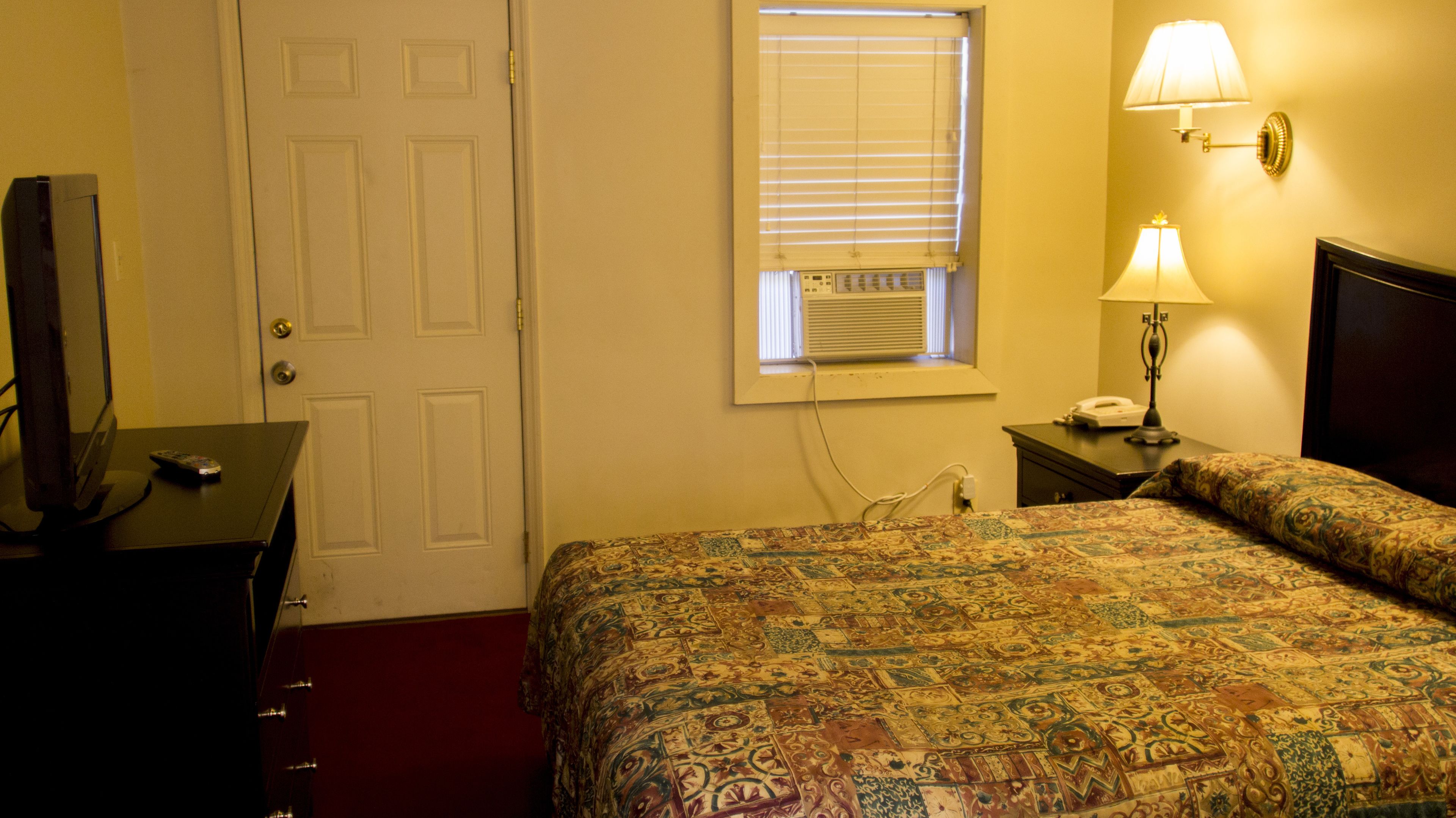 A hotel room with a bed, a desk with a computer and a lamp, a window with blinds, and a door.