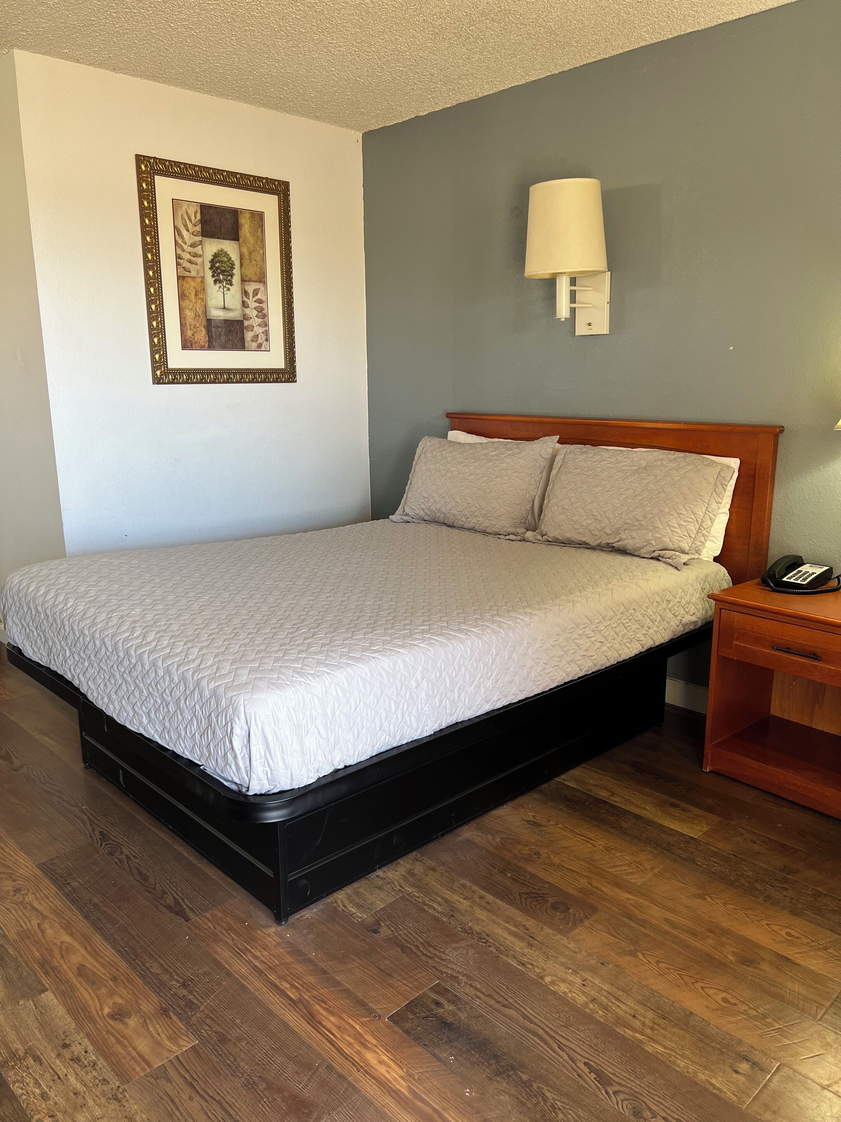 A neatly made bed with a quilted white cover, a wooden headboard, and two pillows. There is a bedside table with a lamp and a phone. The room has wooden flooring and a framed picture on the wall.
