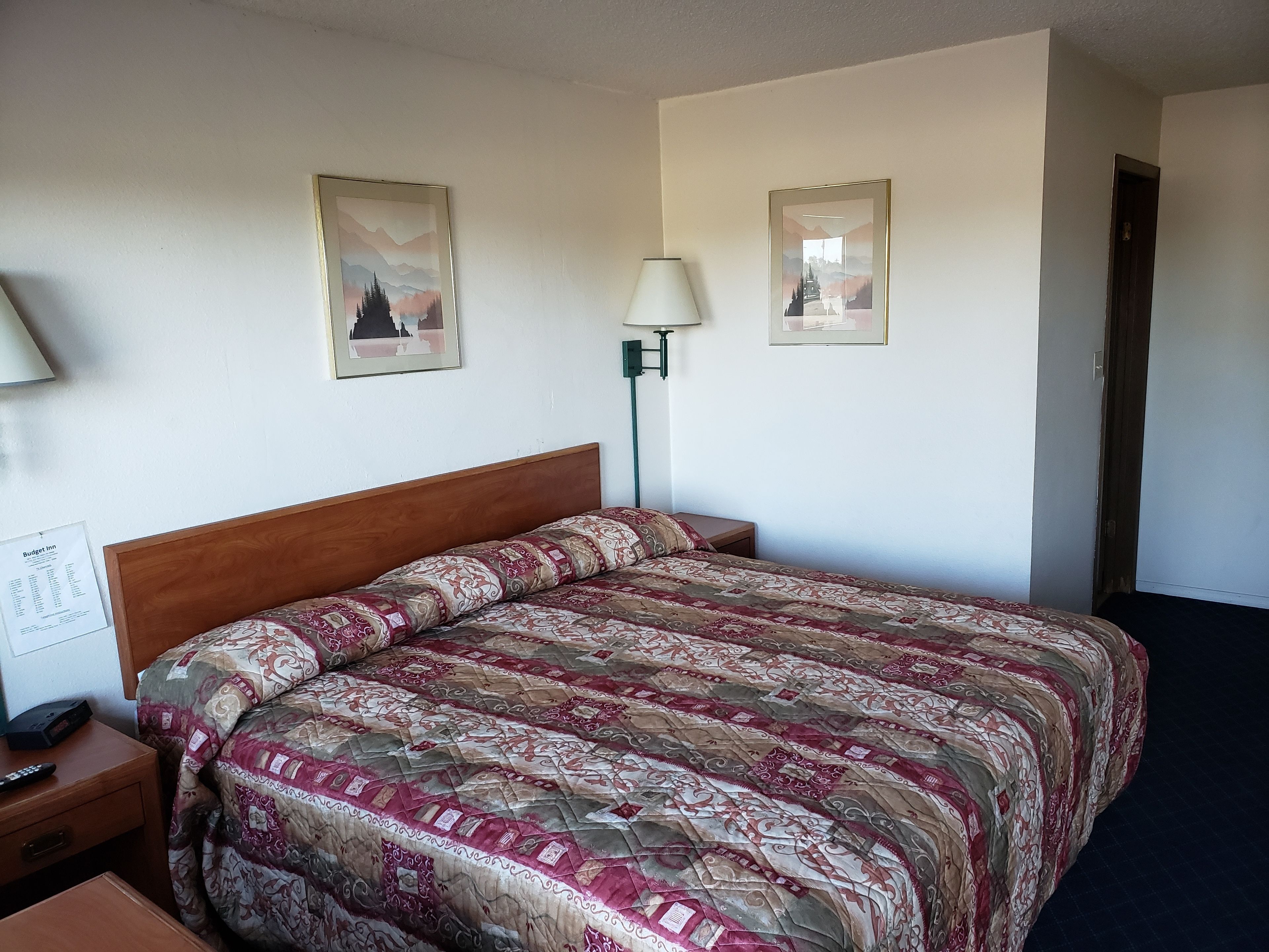 Habitación de hotel con una cama de madera, una mesita de noche, una lámpara y dos cuadros en la pared.
