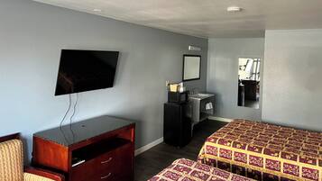 A hotel room with a flat-screen TV mounted on the wall, a wooden dresser with drawers, a bed with a patterned bedspread, a sink, and a mirror.