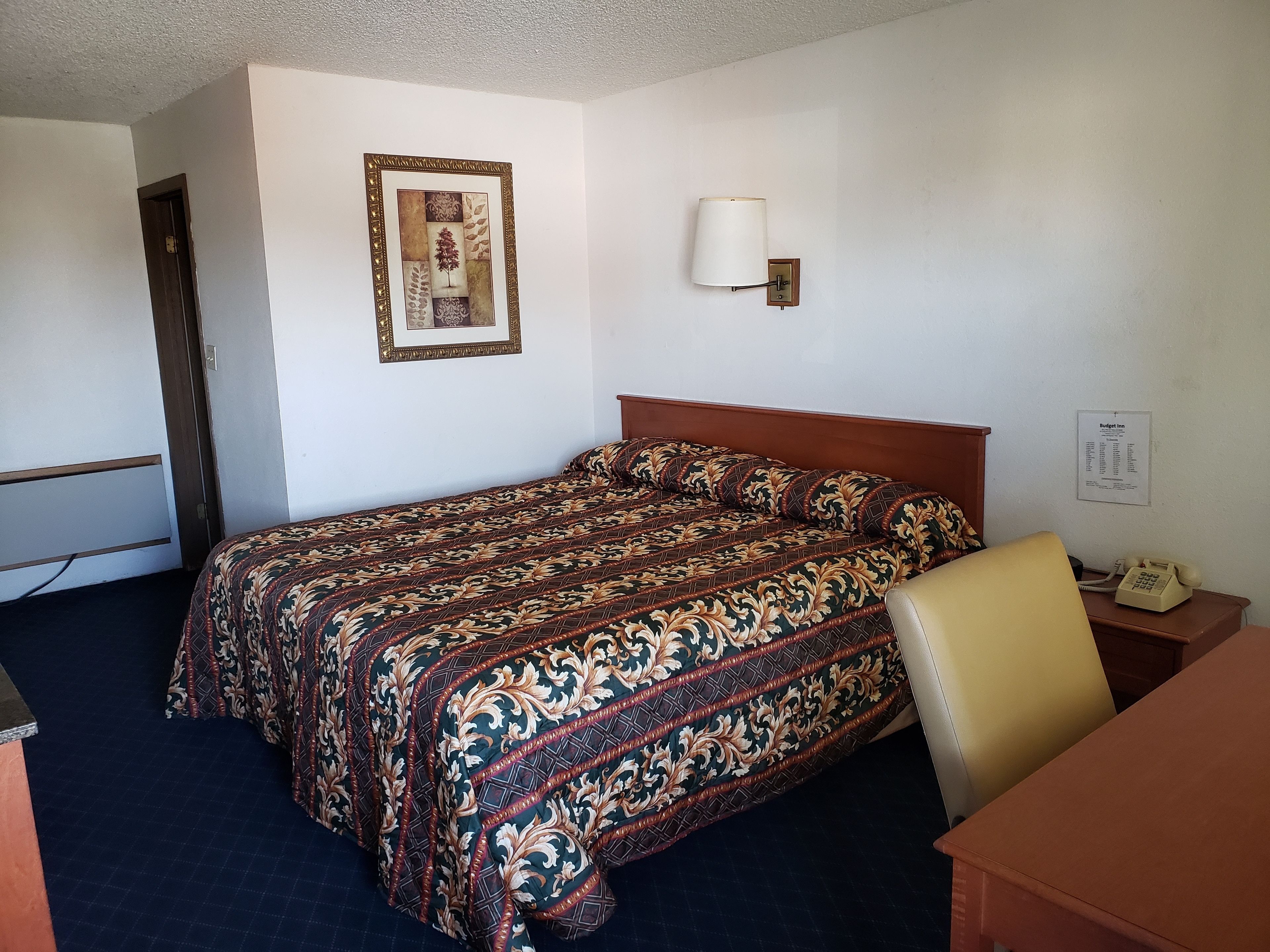 A hotel room with a bed, a desk, a chair, a lamp, and a framed picture on the wall.