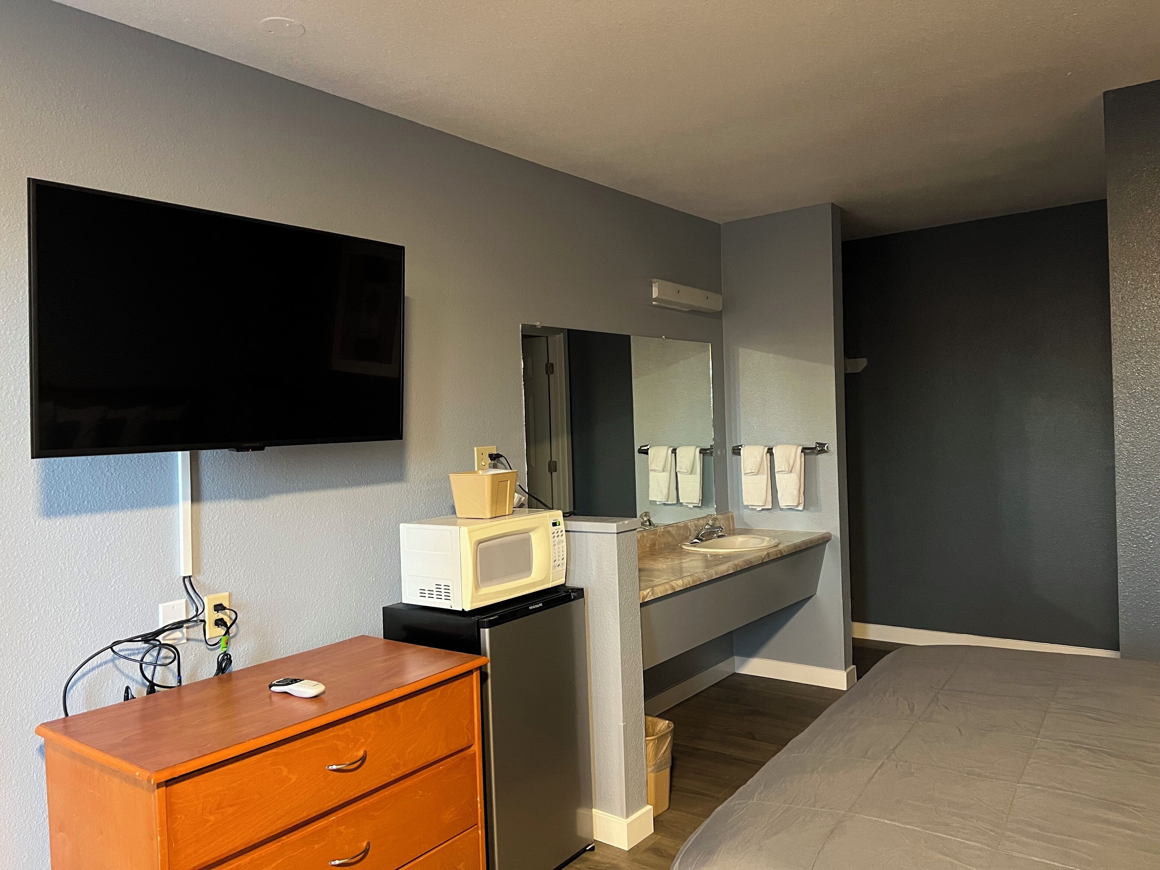 A hotel room with a bed, a wooden dresser, a microwave, a refrigerator, a sink, a mirror, and a television.