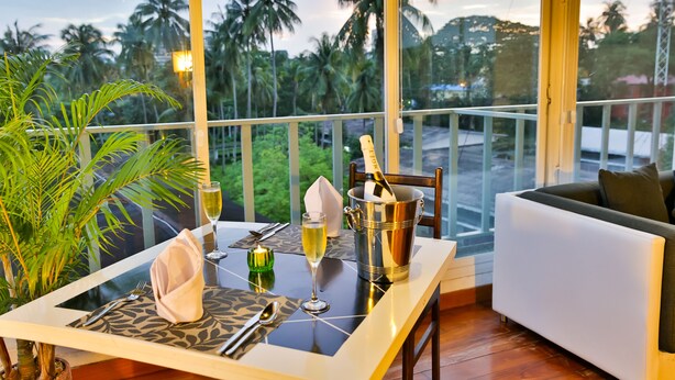 A dining table set with a bottle of champagne, a glass of wine, and a candle, with a view of palm trees and a balcony. - Sky View Hotel Yangon (Yangon)