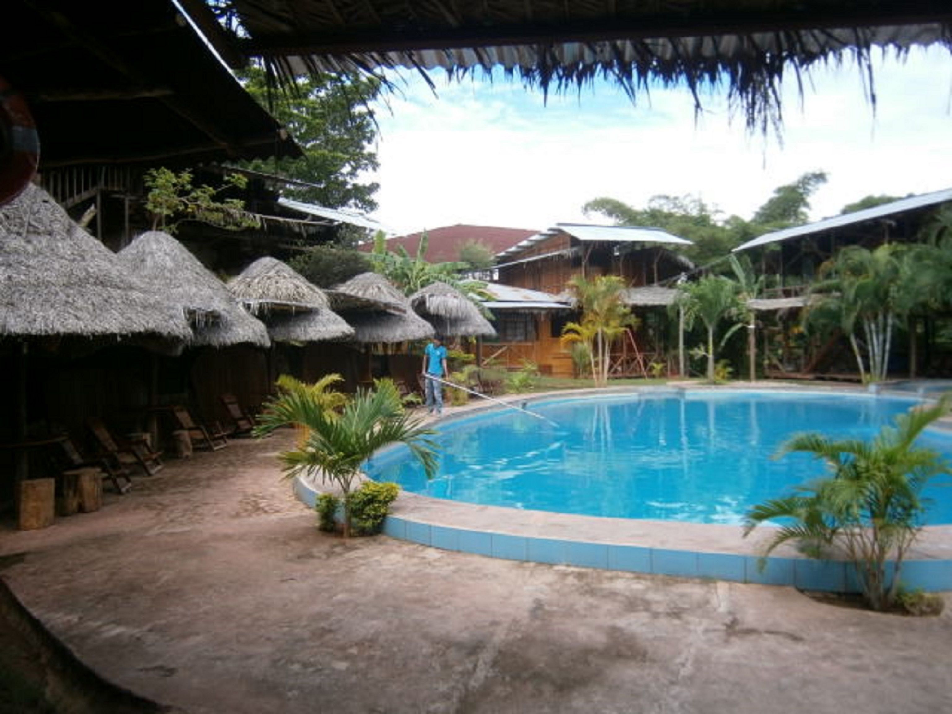 A pool area with a thatched-roof structure, a wooden deck, and a person standing by the pool.
