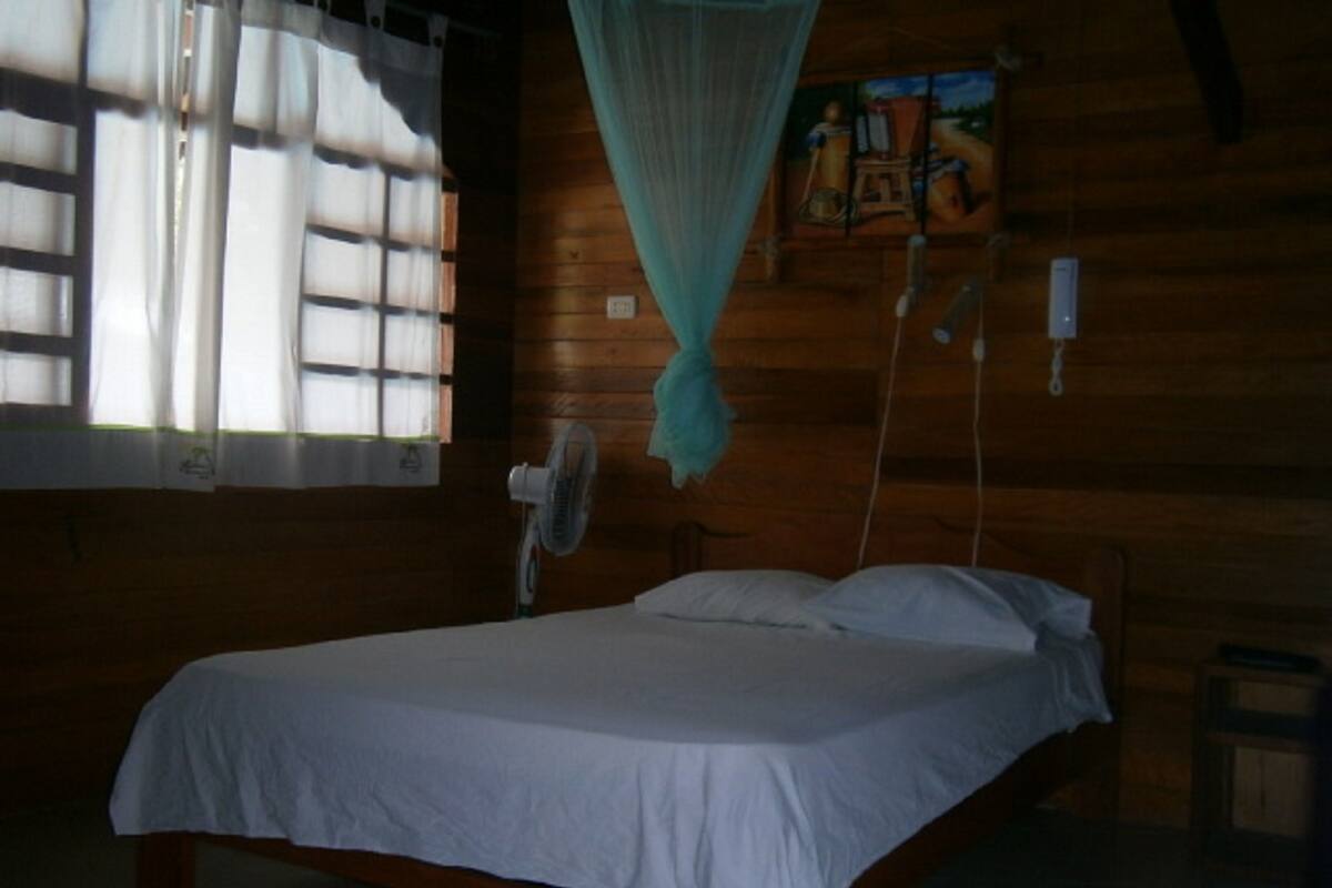 Una habitación de madera con una cama, una ventana con cortinas, un ventilador de pared y una mosquitera colgada.