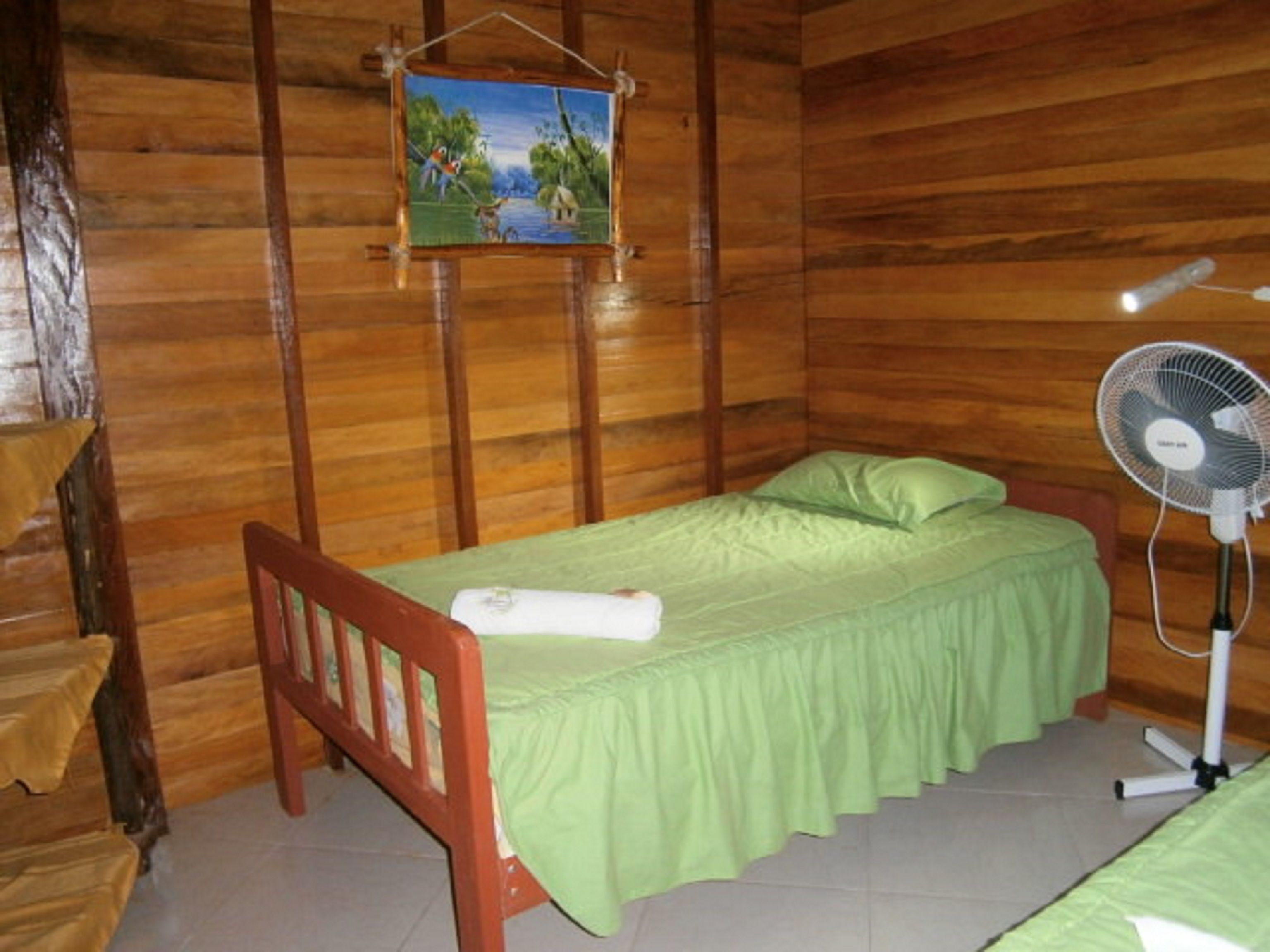 Een houten kamer met een bed, een ventilator en een schilderij aan de muur.