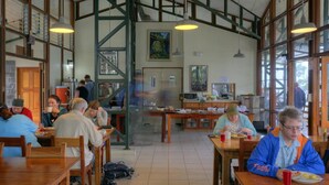 Zona pranzo con tavoli e sedie in legno, una scala e una cucina sullo sfondo.