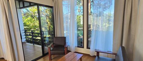 A modern living room with a wooden floor, a sofa, a table, and a chair. Large windows with curtains allow natural light and a view of greenery outside.