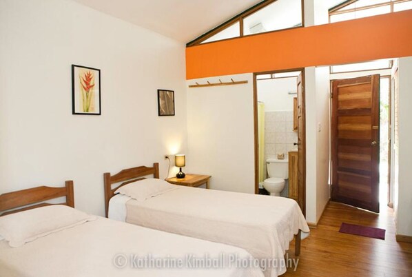 A bedroom with two beds, a nightstand with a lamp, a framed picture on the wall, and a bathroom visible through an open door.