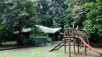 Un terrain de jeux équipé d’un toboggan et d’une structure d’escalade, entouré d’une végétation luxuriante et bordé d’une maison dotée d’un toit végétalisé.