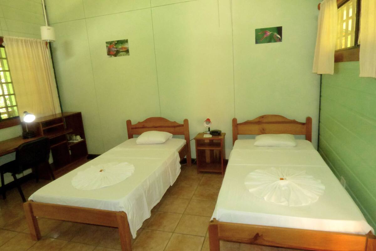 Una habitación con dos camas de madera, cada una con una colcha blanca y un adorno blanco sobre la cama. Hay un escritorio con una silla, una mesita de noche con una lámpara y un estante en la pared.
