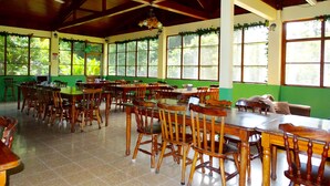 A spacious dining area with wooden tables and chairs, large windows with greenery outside, and a ceiling fan.