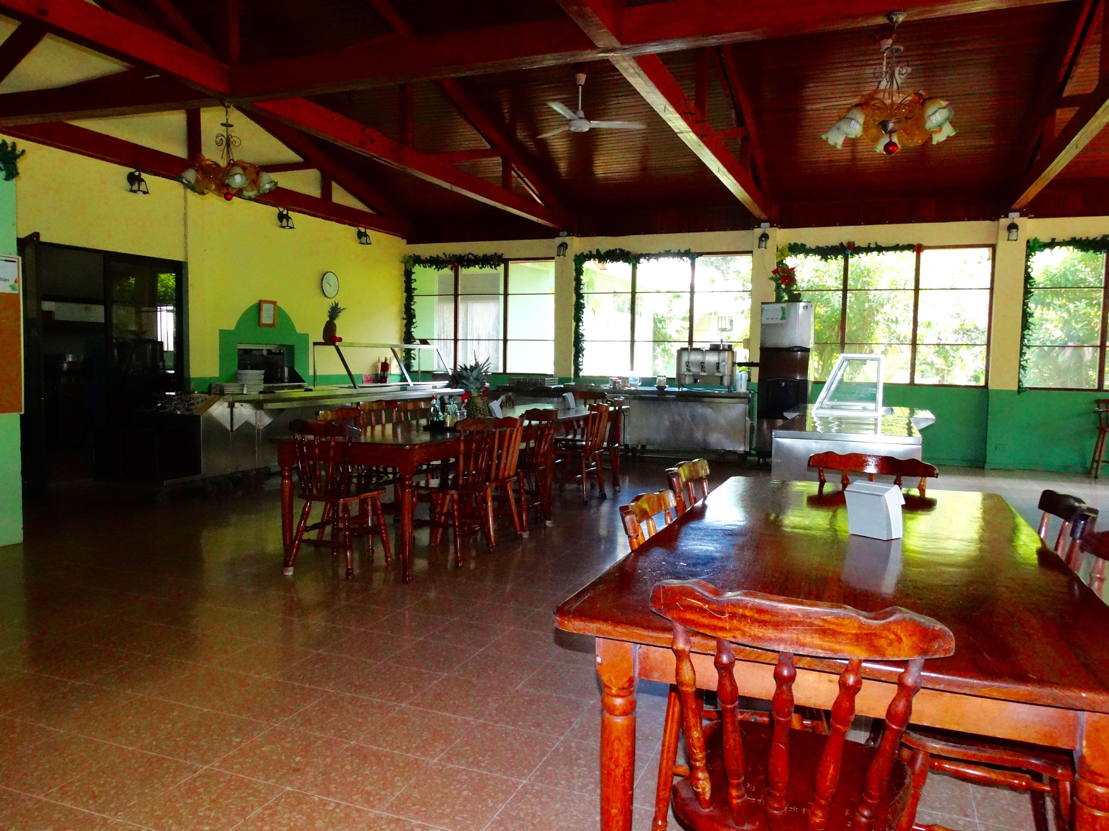 A spacious dining area with wooden tables and chairs, a kitchen in the background, and large windows allowing natural light.