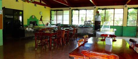 A spacious dining area with wooden tables and chairs, a kitchen in the background, and large windows allowing natural light.