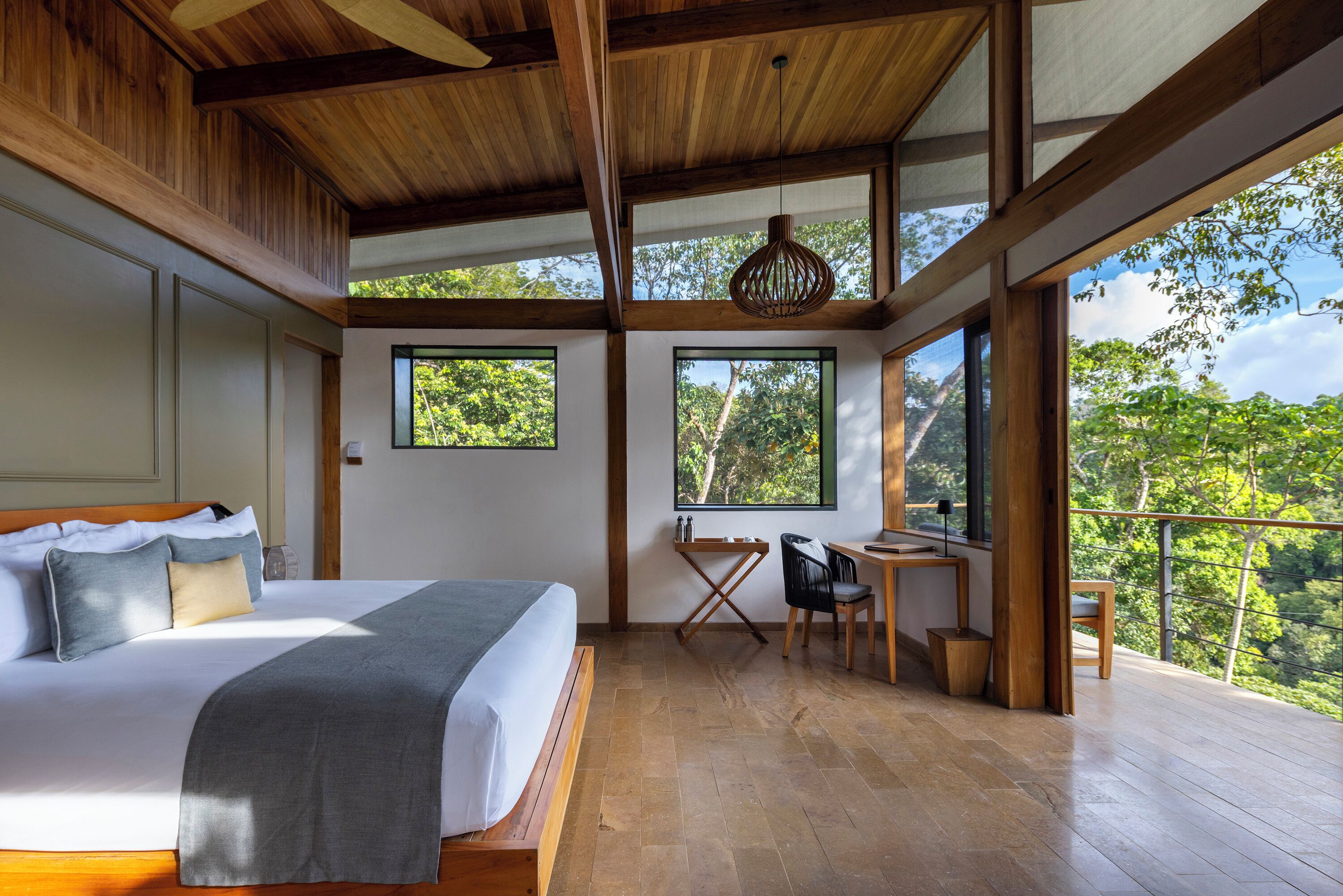 Une chambre avec un grand lit, un bureau et un balcon donnant sur les arbres.