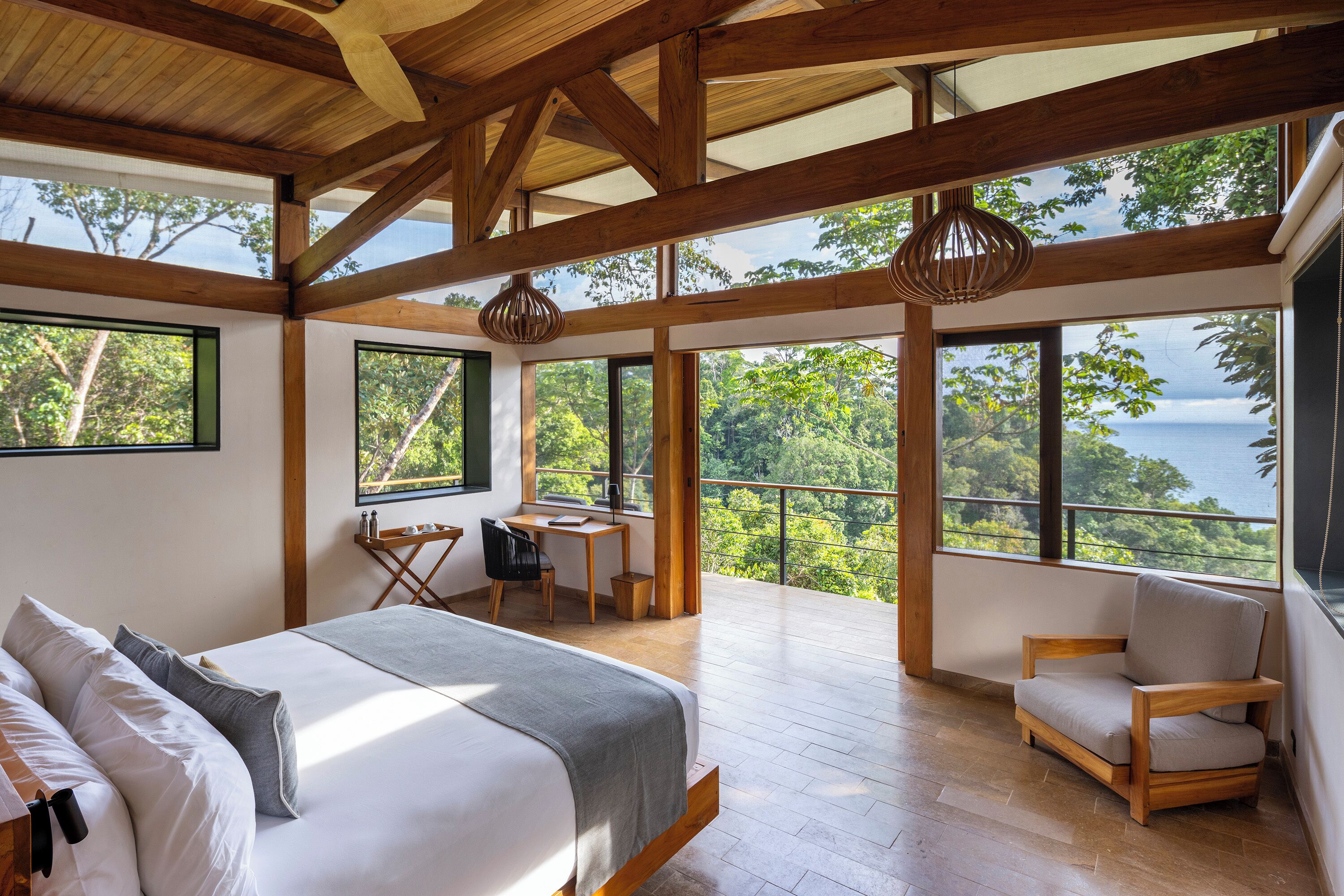 A bedroom with a bed, a desk, and a chair, large windows with a view of trees and a body of water.