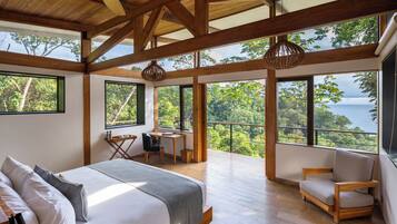 A bedroom with a bed, a desk, and a chair, large windows with a view of trees and a body of water.